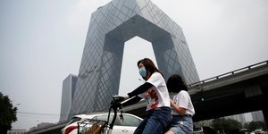 Chine : une femme portant un masque roule en vélo électrique devant le siège de la Télévision centrale de Chine à Pékin le 16 juillet 2020